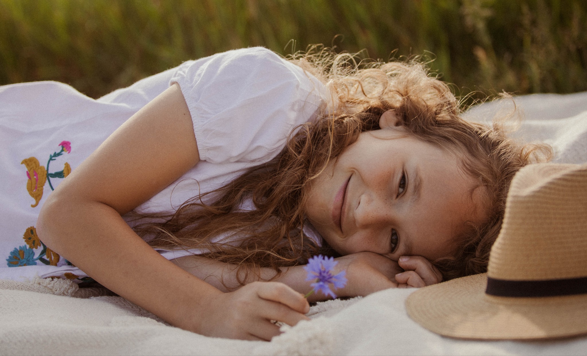 Uśmiechnięta dziewczynka z rudymi kręconymi włosami leży na kocu w trawie, trzymając w ręku niebieski kwiat, obok leży słomkowy kapelusz.