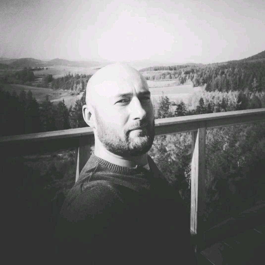 Portret mężczyzny z brodą na tle krajobrazu zalesionych wzgórz, uchwycony w czerni i bieli, z widoczną balustradą.