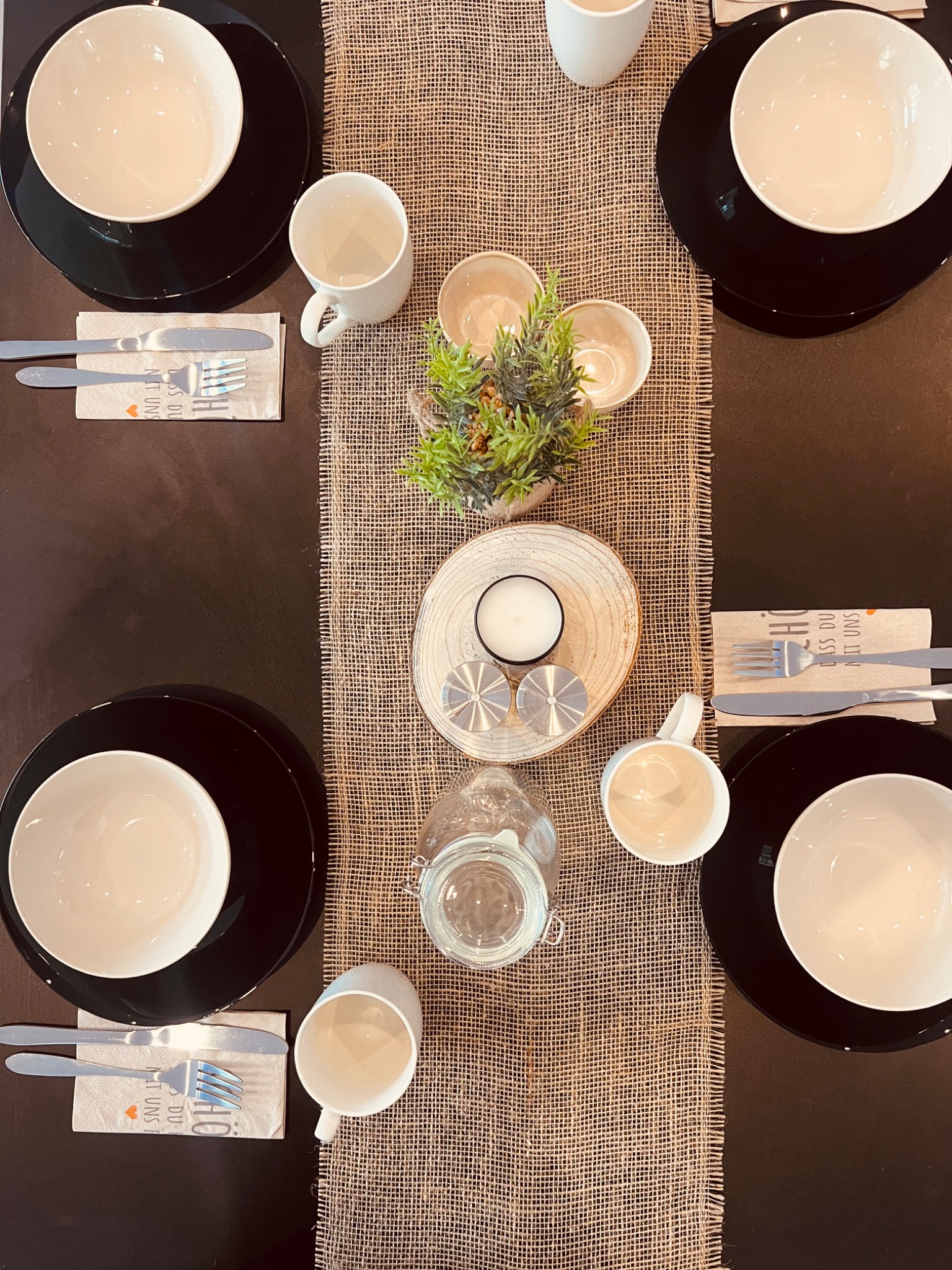 Elegancko nakryty stół z czarnymi talerzami, białymi miseczkami, sztućcami na serwetkach z nadrukiem, białymi kubkami oraz dekoracjami z roślin i świec na bieżniku z juty, ujęcie z góry.