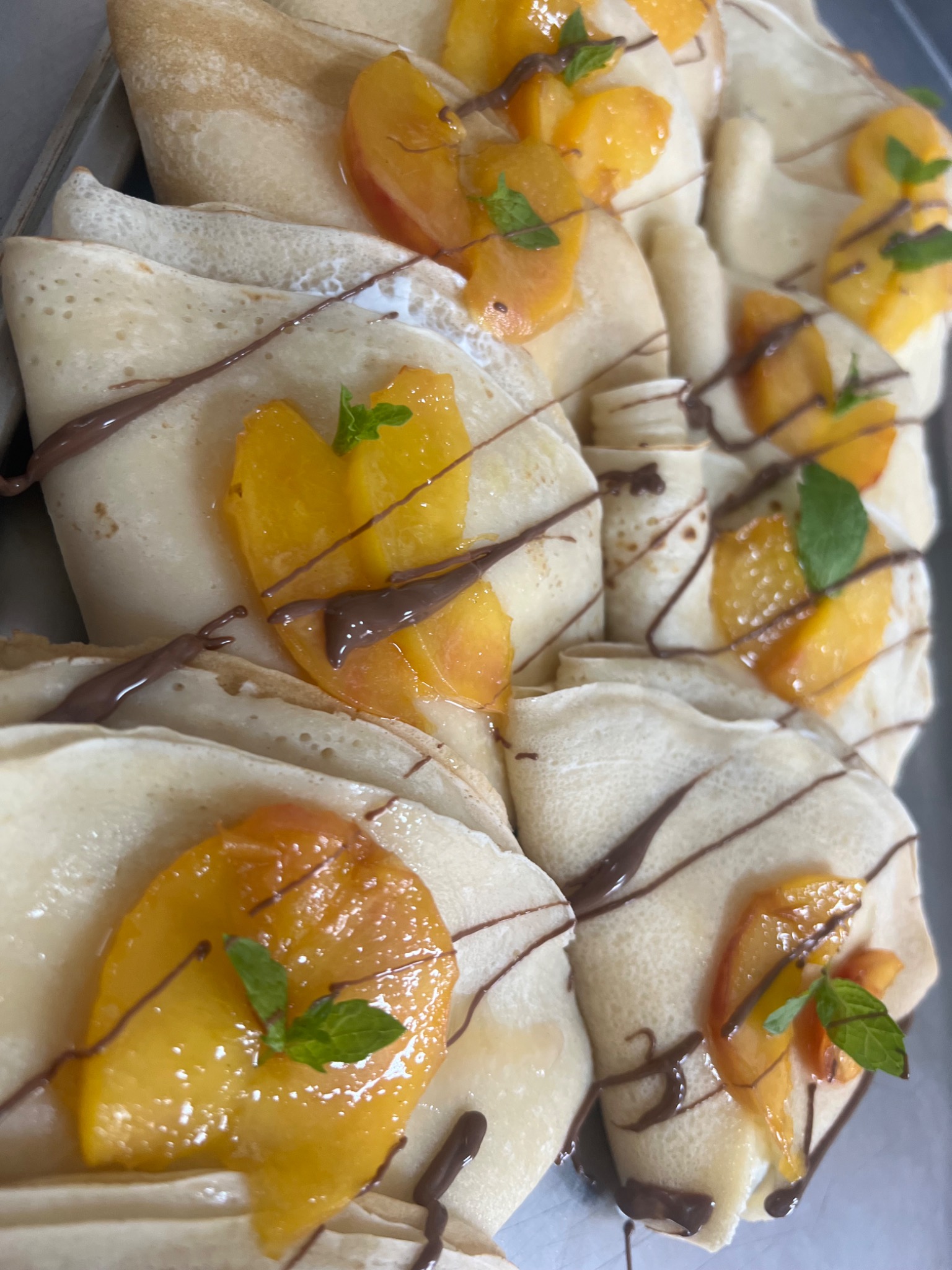 Rząd naleśników zwiniętych w trójkąty, wypełnionych kremem, brzoskwiniami w syropie, polanych czekoladą i udekorowanych listkami mięty, ułożonych na metalowej tacy.