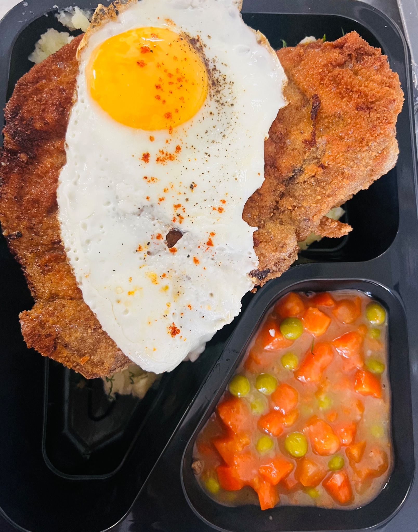 Kotlet schabowy z jajkiem sadzonym i warzywami w sosie, podany w czarnym pudełku na wynos.