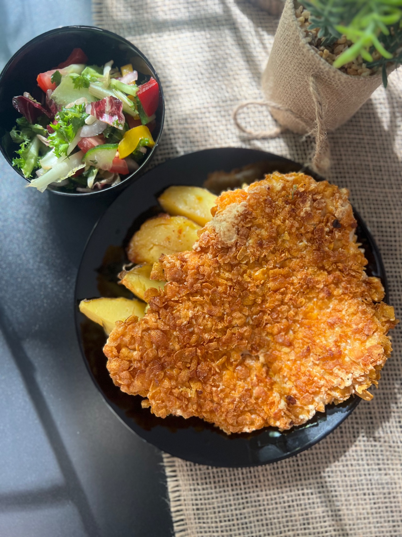 Gotowy obiad: panierowany kotlet w płatkach kukurydzianych, pieczone ziemniaki i sałatka warzywna w czarnej miseczce, podane na czarnym talerzu, obok dekoracyjna roślina w jutowym worku.
