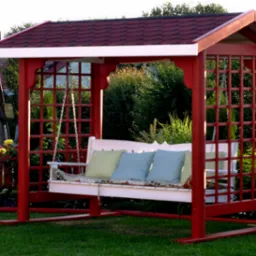 Czerwona, drewniana huśtawka ogrodowa z kratowanymi bokami i dachem z czerwonej dachówki, z białym siedziskiem i poduszkami, otoczona zielenią i kwiatami, w tle dom.