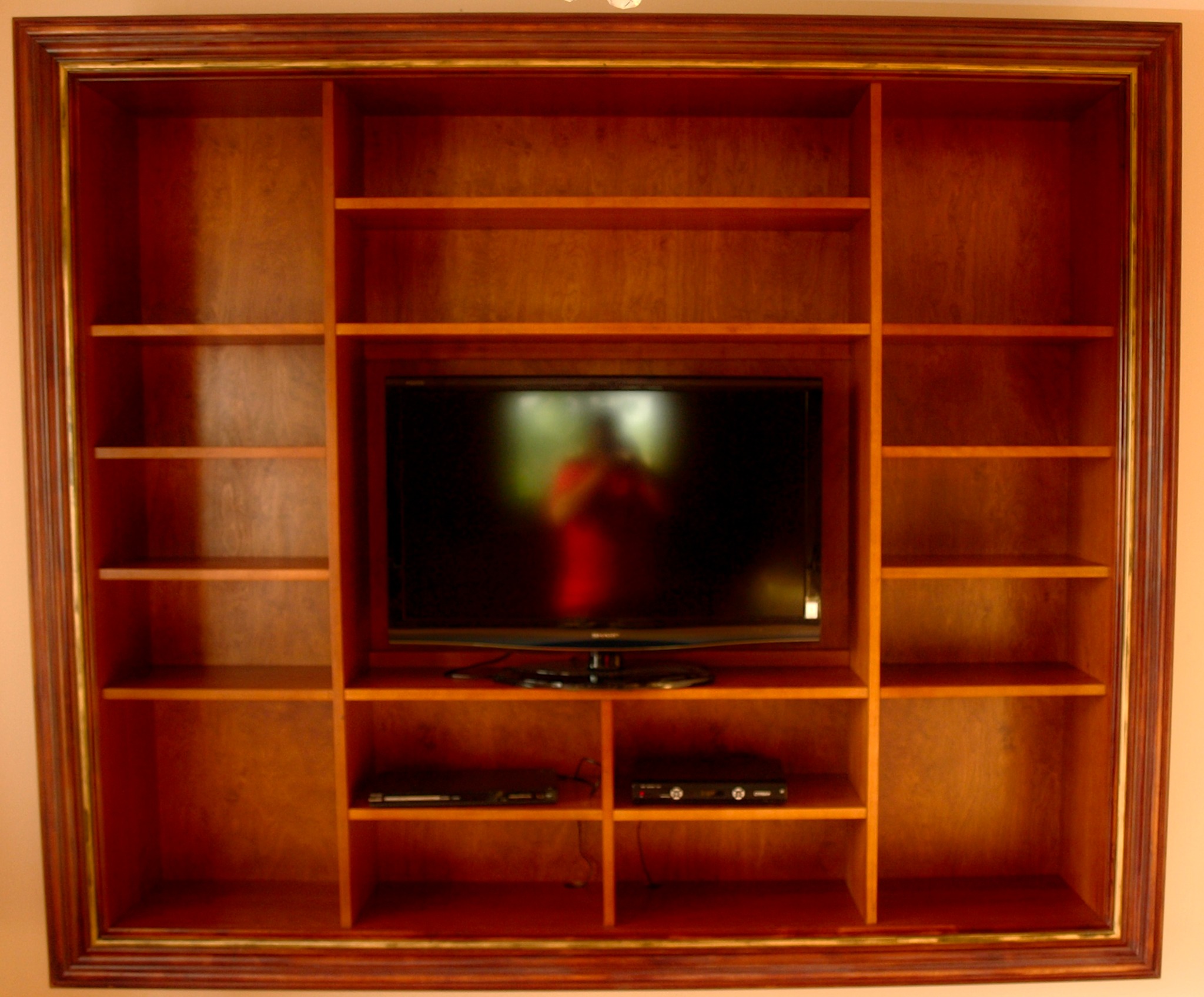 Drewniana szafa na wymiar w zabudowie ściennej z miejscem na telewizor i sprzęt audio-wideo, wykończona w ciepłym odcieniu brązu z dekoracyjną ramą.