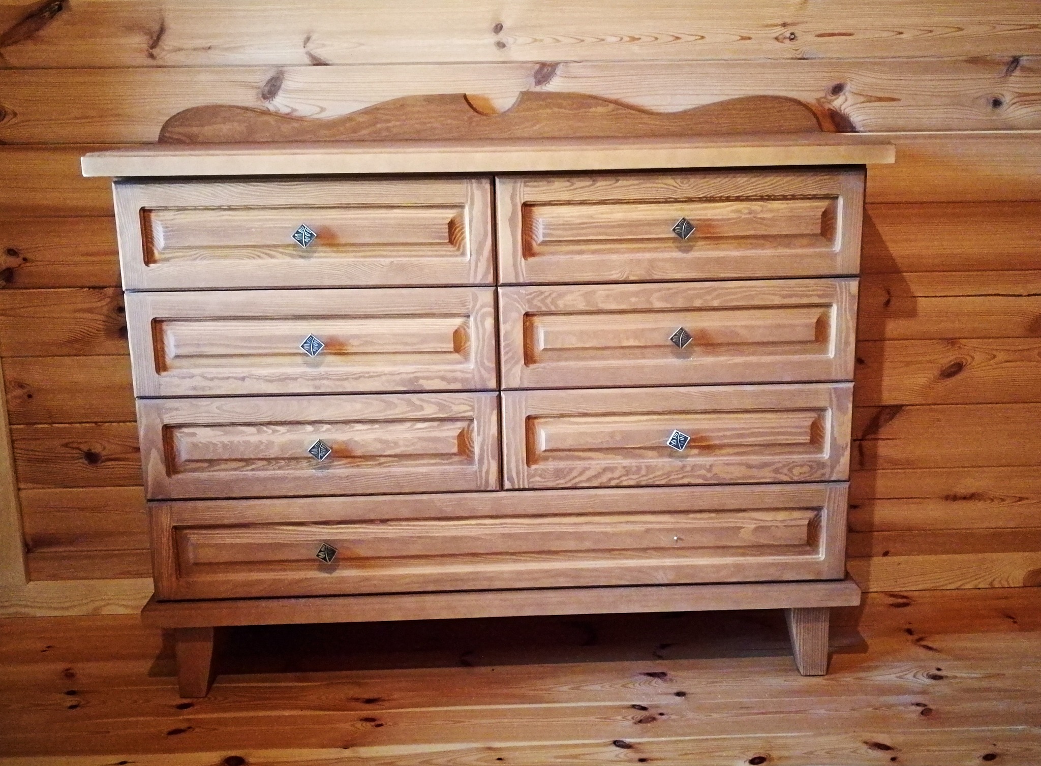 Drewniana komoda z sześcioma szufladami i ozdobnymi gałkami w kształcie rombów, na tle ściany i podłogi z desek w podobnym odcieniu.