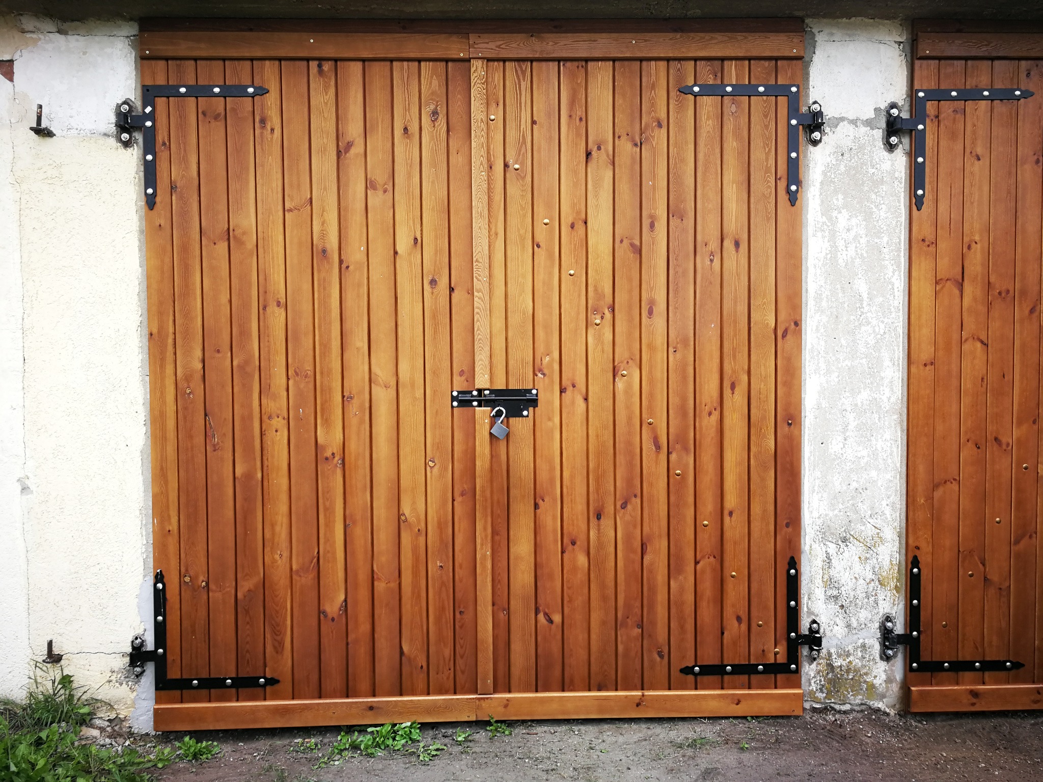 Dwuskrzydłowe, drewniane drzwi garażowe w kolorze miodowym, z czarnymi, zdobionymi zawiasami i zamkiem na środku, umieszczone w betonowej ramie.