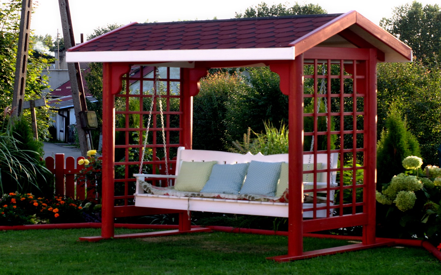 Czerwona, drewniana huśtawka ogrodowa z kratowanymi bokami i dachem z czerwonej dachówki, z białym siedziskiem i poduszkami, otoczona zielenią i kwiatami, w tle dom.
