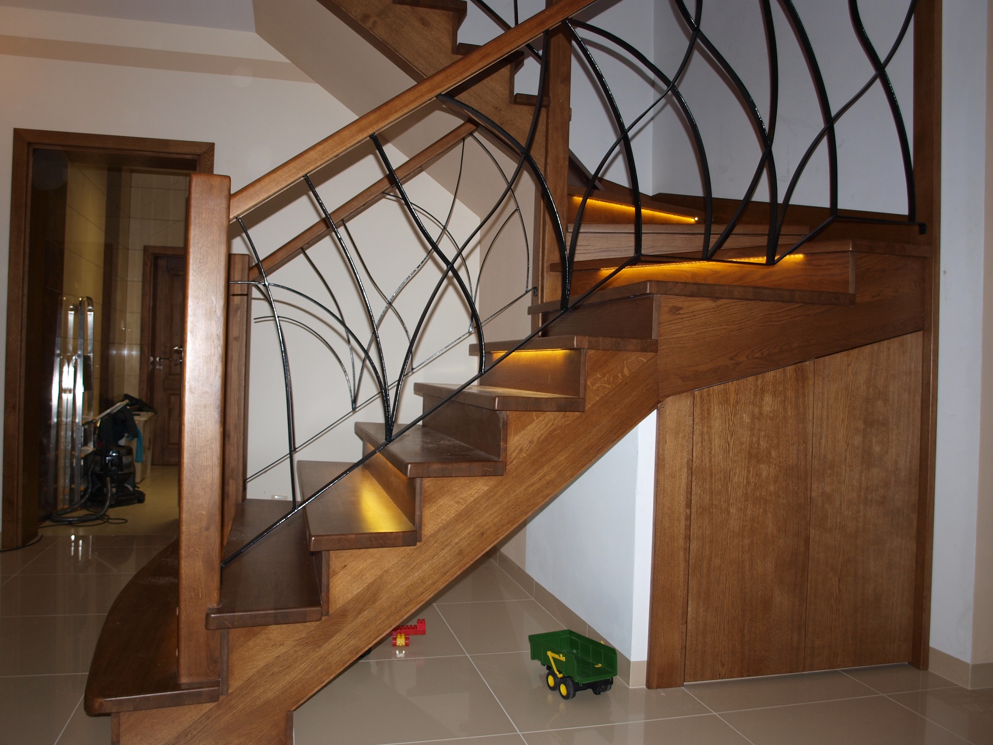 Drewniane schody z oświetleniem LED pod stopniami i ozdobną, metalową balustradą. Na podłodze leżą dziecięce zabawki.