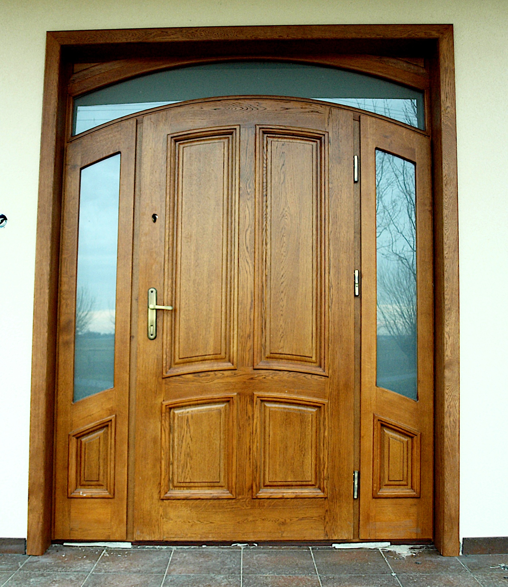 Drewniane drzwi wejściowe z szybami po bokach i nad górną krawędzią, widoczne z zewnątrz na tle białej ściany i brązowej podłogi z płytek.