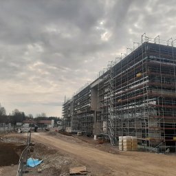 Continum sp. z o. o. - Wielopiętrowy budynek w trakcie budowy, otoczony rusztowaniami, z widoczną konstrukcją żelbetową i stertą materiałów budowlanych na pierwszym planie, na tle pochmurnego nieba.