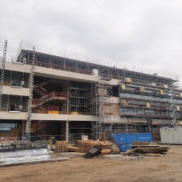 Continum sp. z o. o. - Wielopiętrowy budynek w trakcie budowy, otoczony rusztowaniami, z widocznymi schodami wewnętrznymi i kontenerami budowlanymi na placu. Na pierwszym planie materiały budowlane i palety.