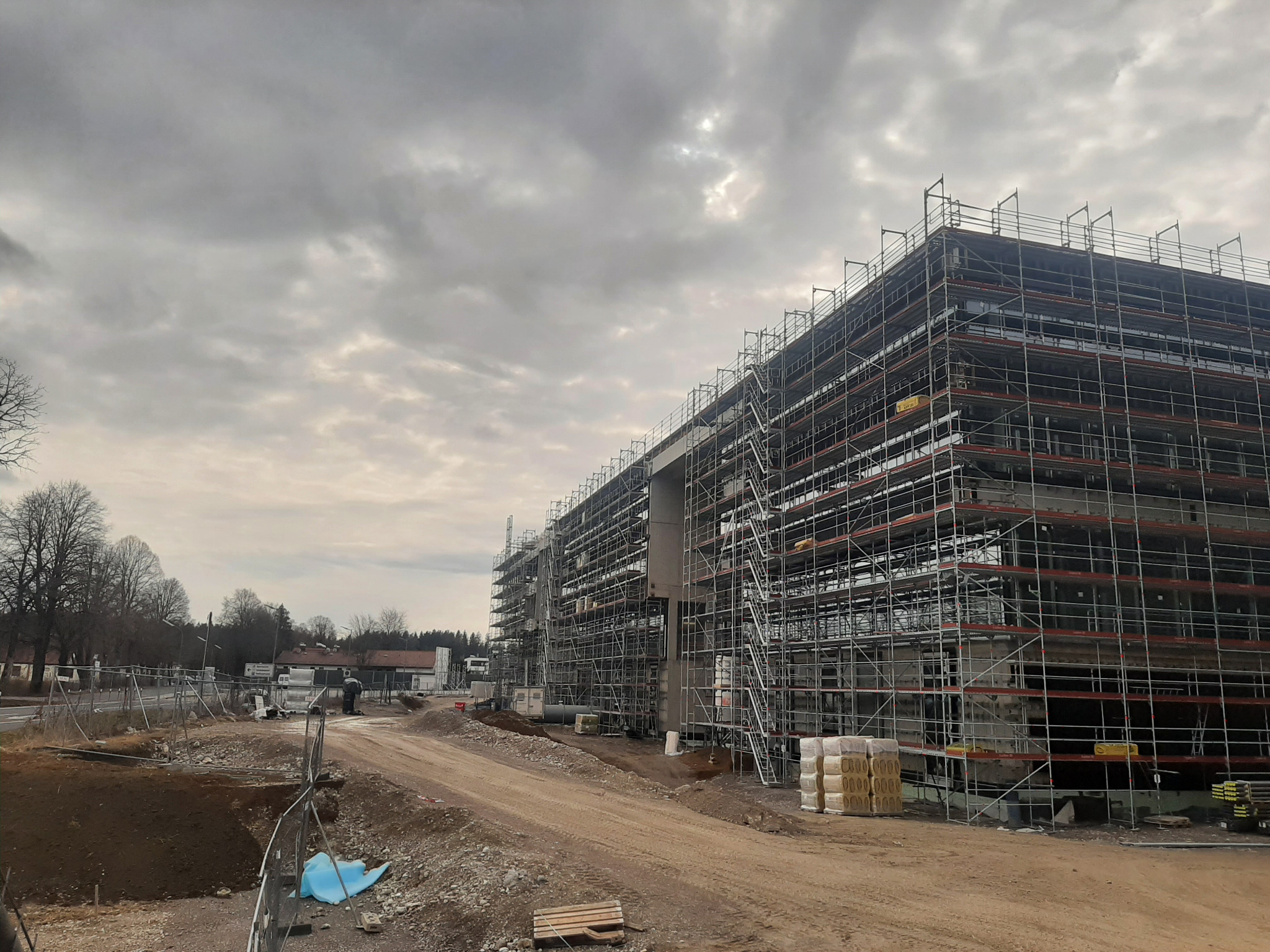 Wielopiętrowy budynek w trakcie budowy, otoczony rusztowaniami, z widoczną konstrukcją żelbetową i stertą materiałów budowlanych na pierwszym planie, na tle pochmurnego nieba.