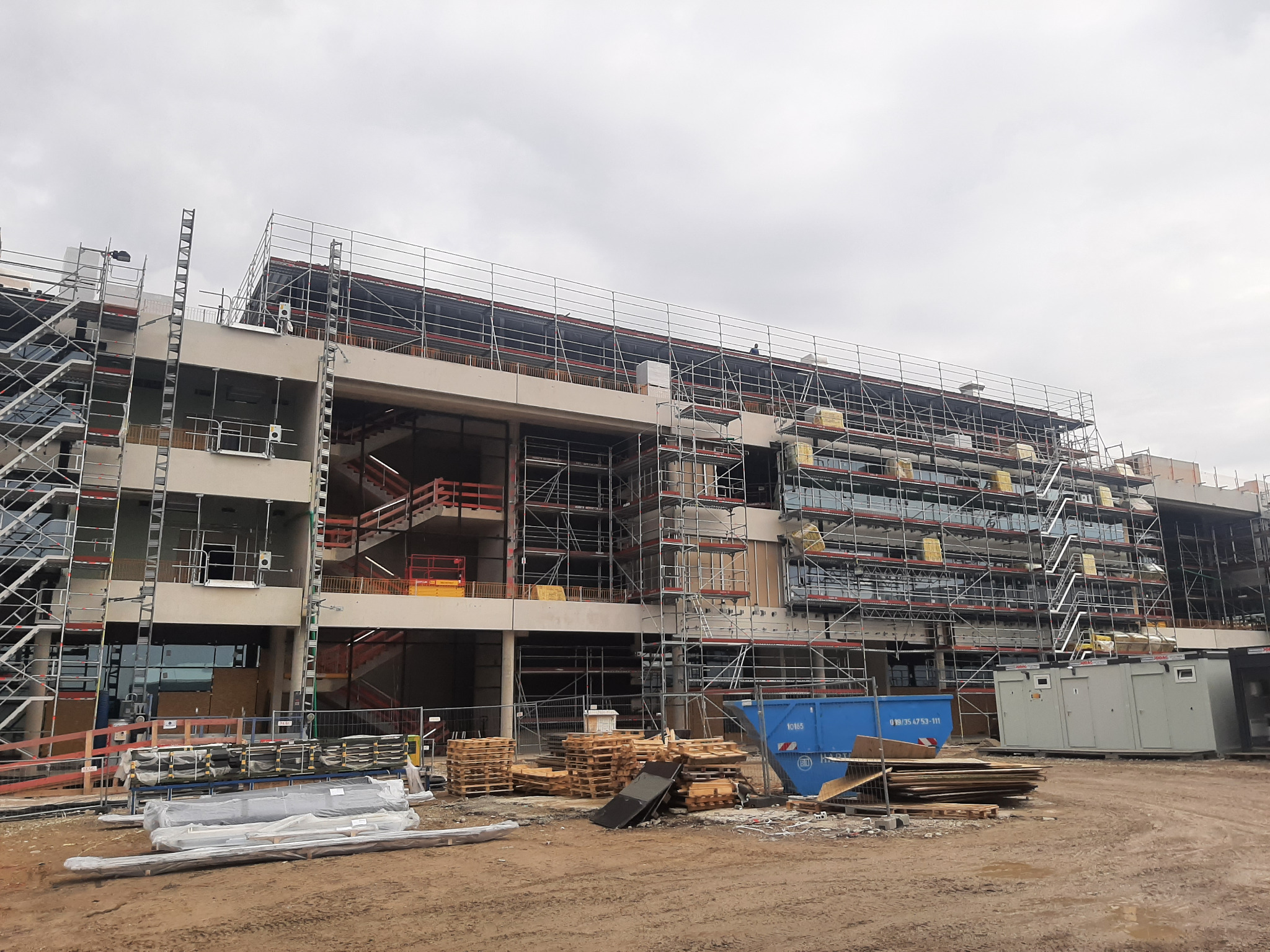 Wielopiętrowy budynek w trakcie budowy, otoczony rusztowaniami, z widocznymi schodami wewnętrznymi i kontenerami budowlanymi na placu. Na pierwszym planie materiały budowlane i palety.