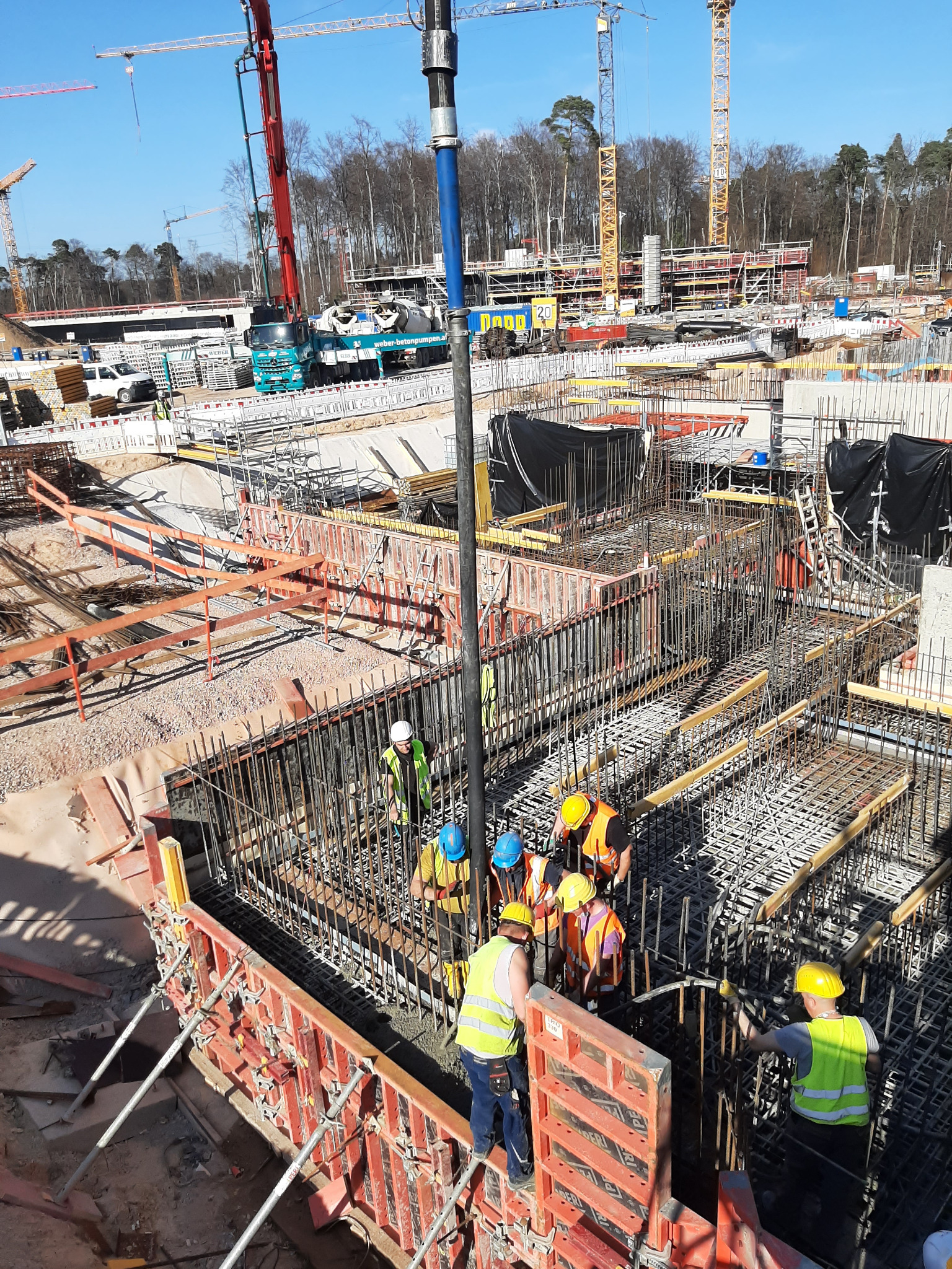 Wysoki kąt widzenia na plac budowy z pracownikami w kaskach i kamizelkach odblaskowych w trakcie wylewania betonu do szalunków zbrojeniowych, w tle widoczny mikser do betonu i żurawie budowlane.