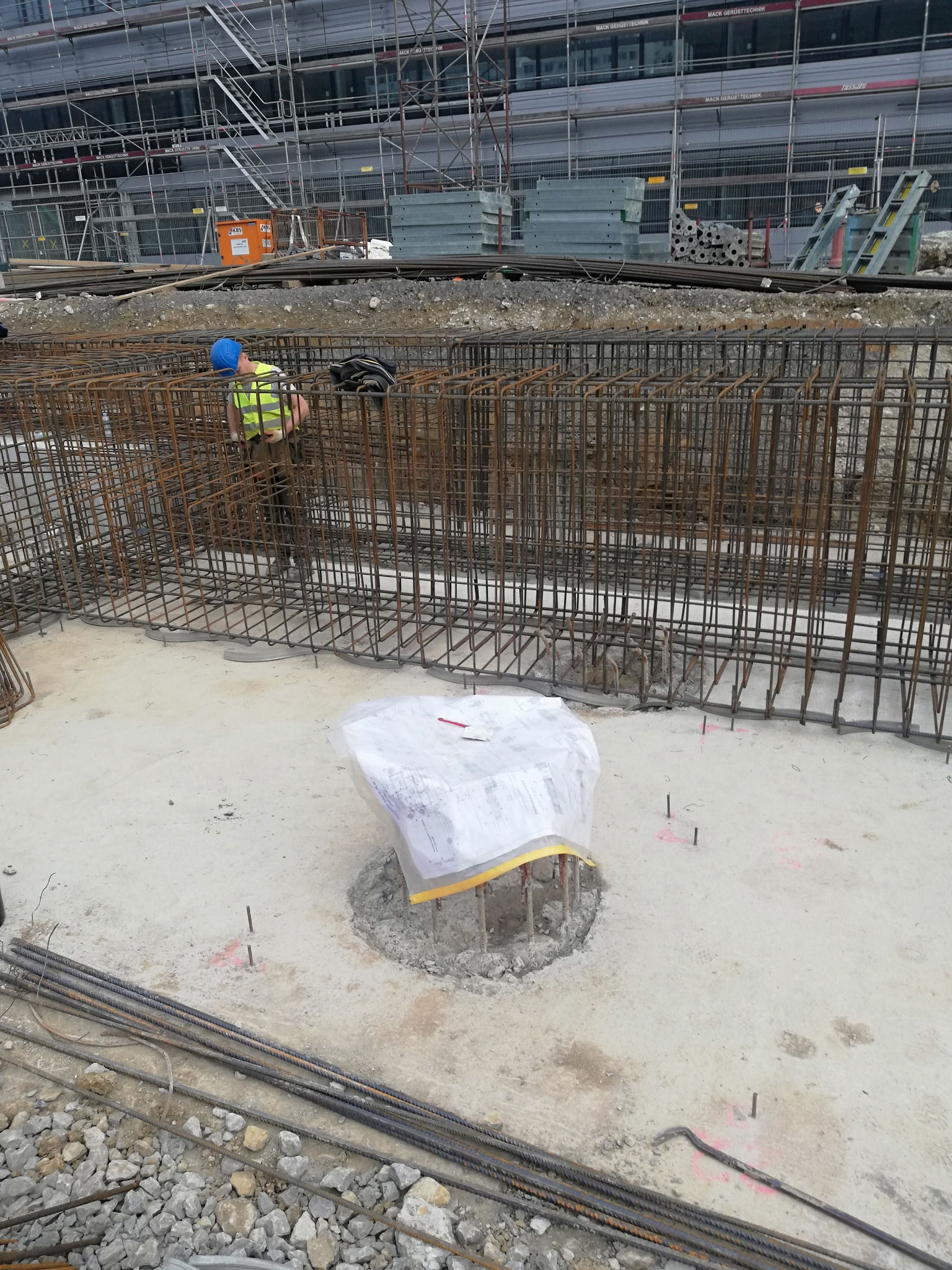 Pracownik w kasku i kamizelce odblaskowej przy zbrojeniu fundamentu, widoczne pręty zbrojeniowe, betonowa podstawa owinięta folią i planami, w tle rusztowania i elementy konstrukcyjne budynku.