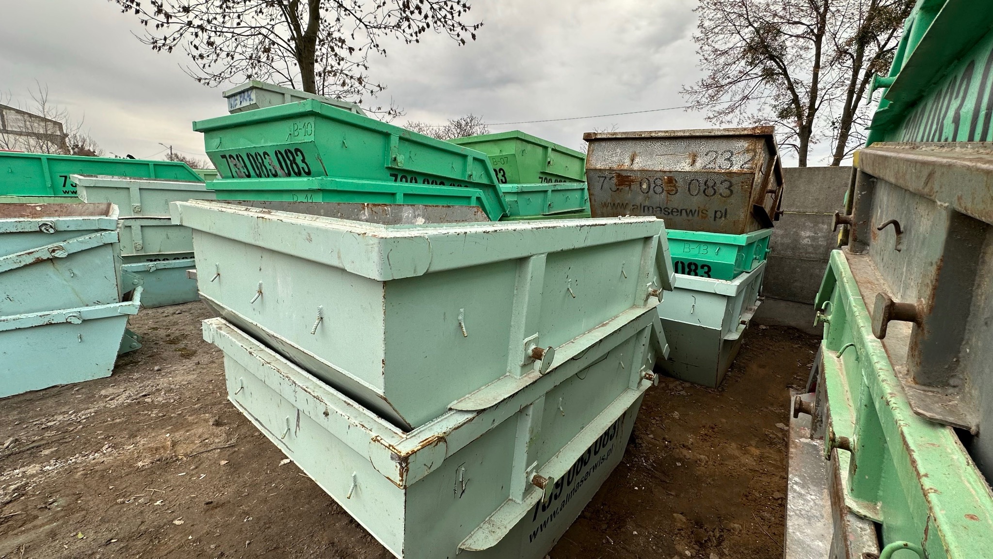 Stos zielonych i szarych kontenerów z widocznymi numerami telefonów i adresami stron internetowych, ustawionych na ziemi pod pochmurnym niebem.