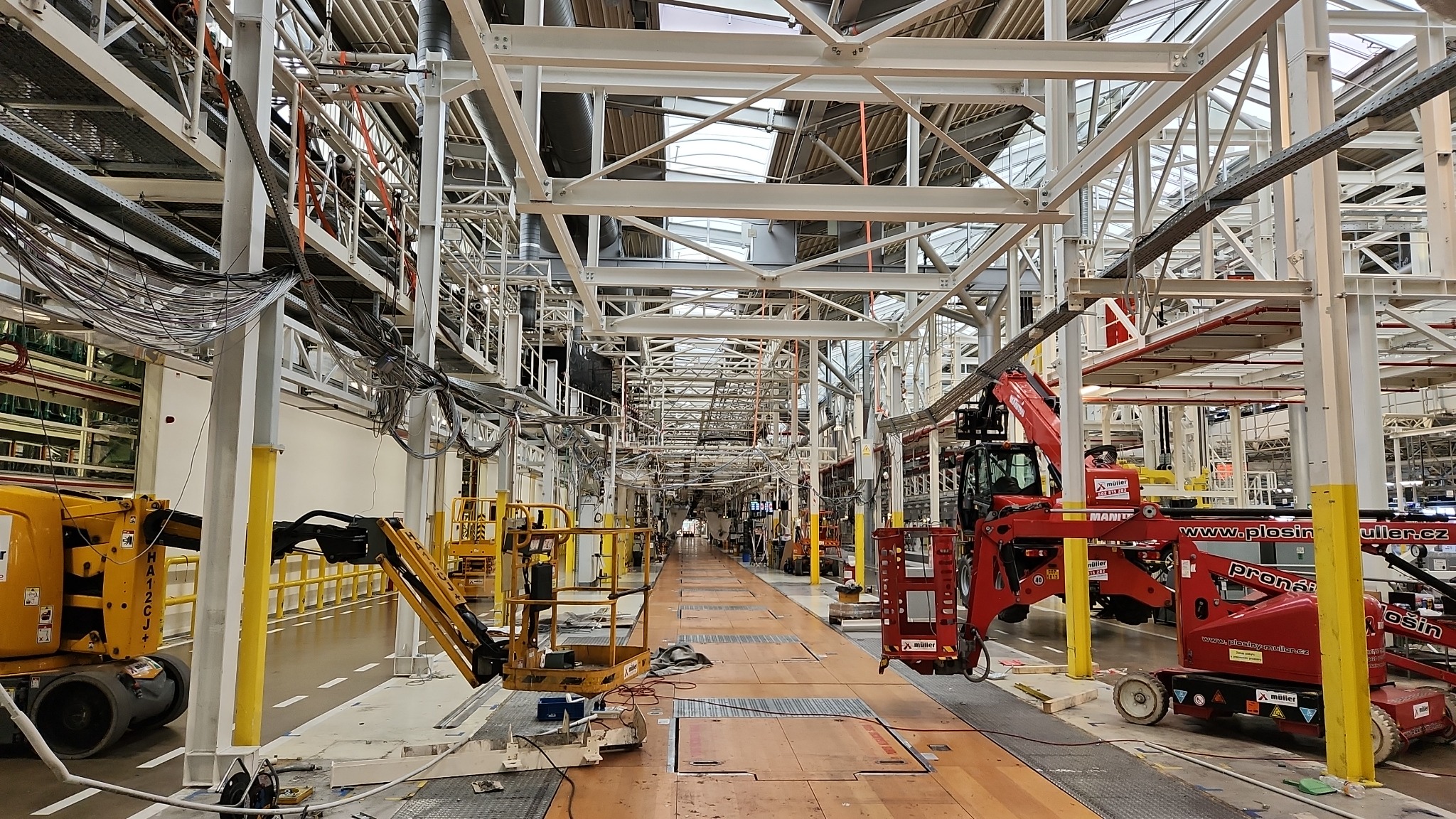 Wnętrze hali w trakcie budowy, widoczne stalowe konstrukcje, podnośniki koszowe i liczne przewody zwisające z sufitu. Jasne oświetlenie dzienne wpada przez okna dachowe.