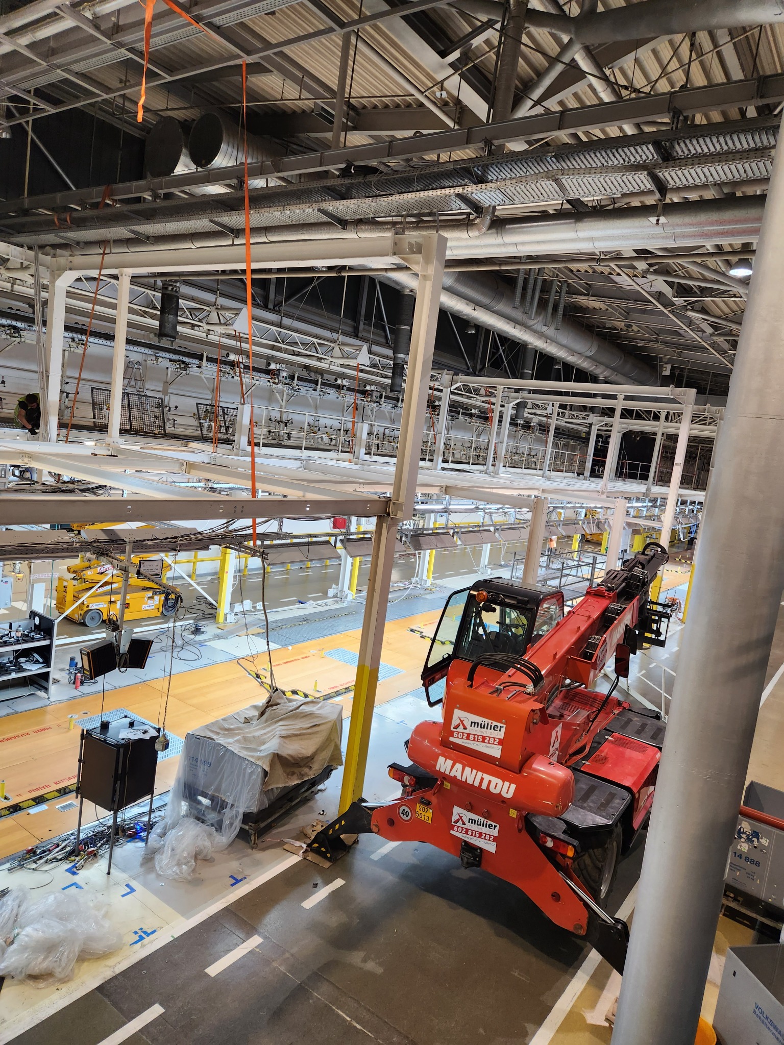 Wnętrze hali produkcyjnej w trakcie budowy, widok z góry na czerwoną ładowarkę teleskopową Manitou, z widocznymi elementami konstrukcji stalowej i instalacji pod sufitem.