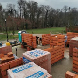 Wznoszenie ścian domu z bloczków ceramicznych Leier Porotherm, widoczny pracownik przy betoniarce, palety bloczków, przenośna toaleta w tle, pochmurne niebo.