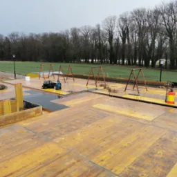 Przygotowanie płyty fundamentowej z widocznym zbrojeniem, deskowaniem i sprzętem budowlanym na tle zieleni w pochmurny dzień.