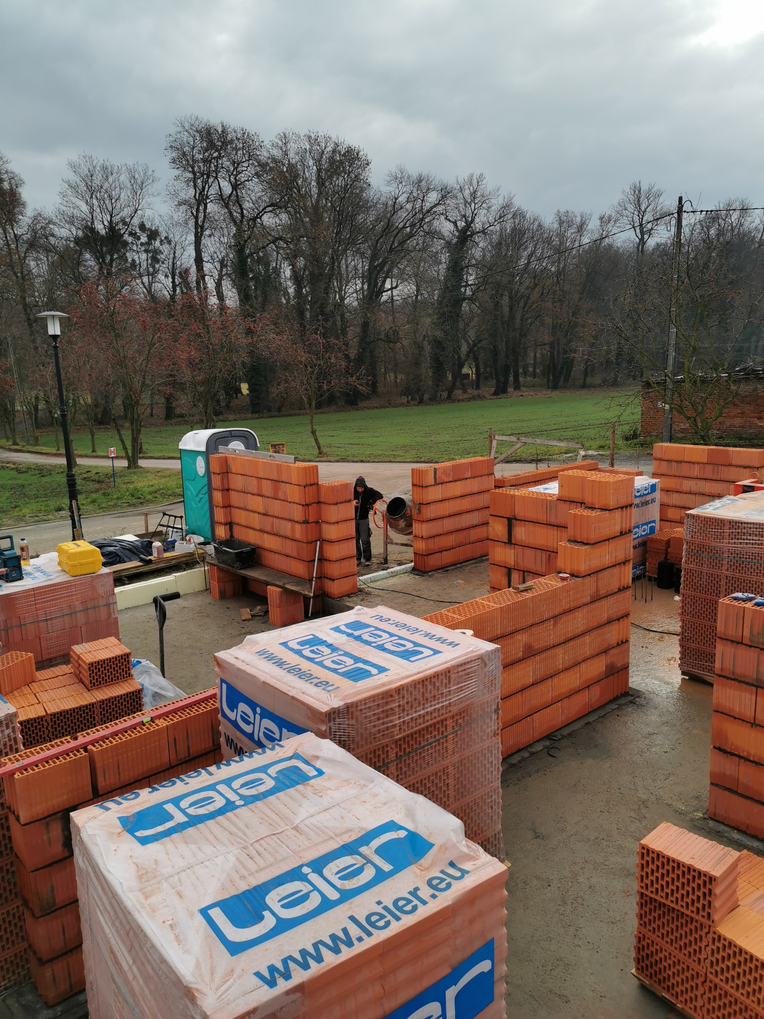 Wznoszenie ścian domu z bloczków ceramicznych Leier Porotherm, widoczny pracownik przy betoniarce, palety bloczków, przenośna toaleta w tle, pochmurne niebo.