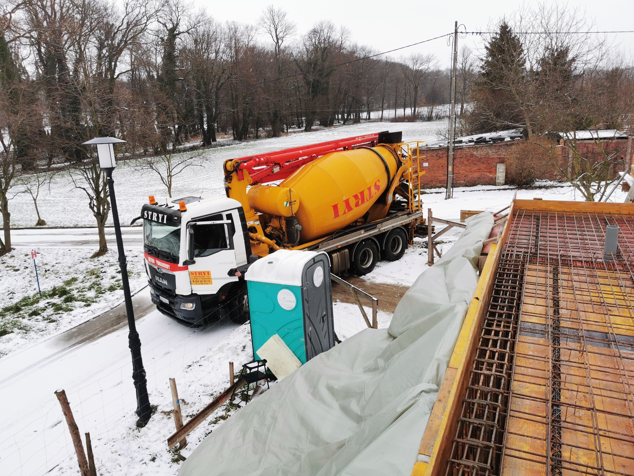 Żółta betoniarka firmy STRYI na budowie w zimowy dzień, widoczne szalunki i zbrojenie fundamentów, przenośna toaleta Toi Toi.