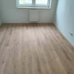 Jasna podłoga winylowa imitująca dąb w pustym, pomalowanym na biało pokoju z grzejnikiem pod oknem.