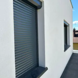 Antracytowa, adaptacyjna osłona okienna z lamelami PA39, zamontowana na elewacji budynku o jasnym tynku strukturalnym, z widocznym fragmentem okna i niebieskim niebem w tle.