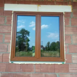 Nowe dwuskrzydłowe okno w kolorze złoty dąb, zamontowane w surowej ścianie z czerwonej cegły, z widoczną pianką montażową i odbiciem krajobrazu w szybach.