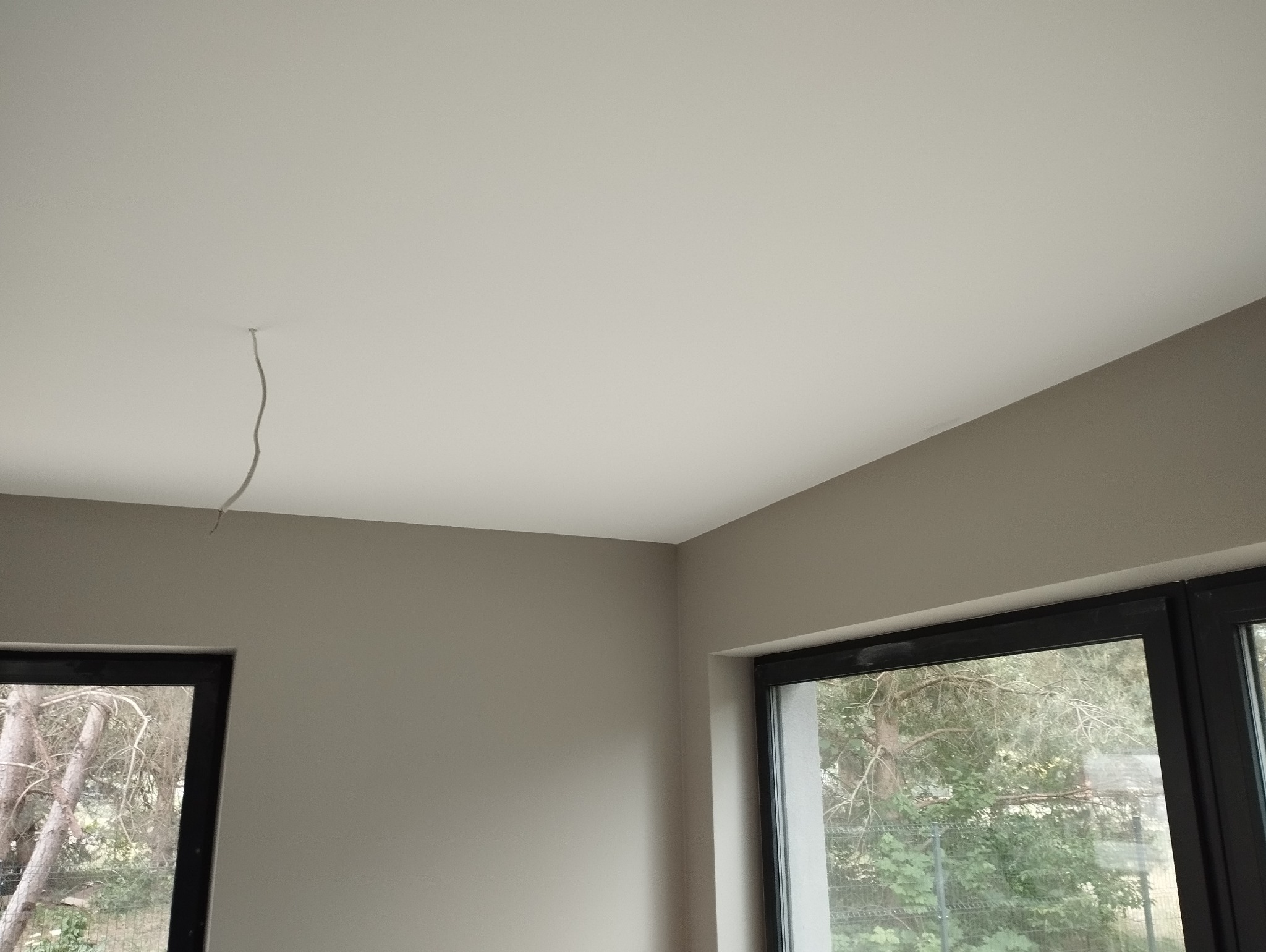 Świeżo pomalowane na szaro ściany i biały sufit w pokoju w Warszawie, widok na zewnątrz przez czarne ramy okienne, widoczny wystający kabel ze ściany.