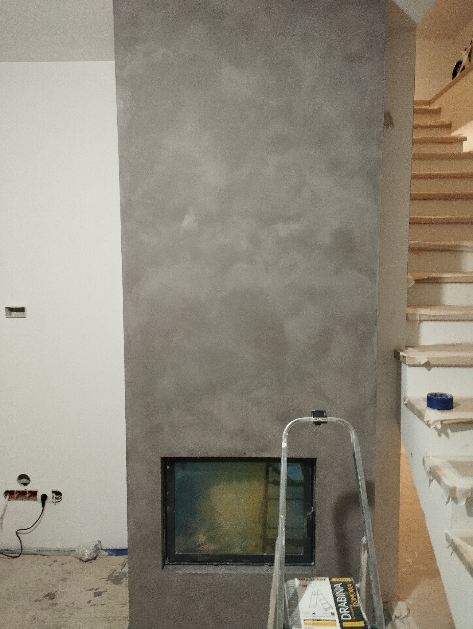 Ściana z efektem dekoracyjnym w odcieniach szarości, wbudowany w nią czarny wkład kominkowy, drabina oparta o ścianę, widoczne schody w tle, surowy stan wykończenia podłogi.