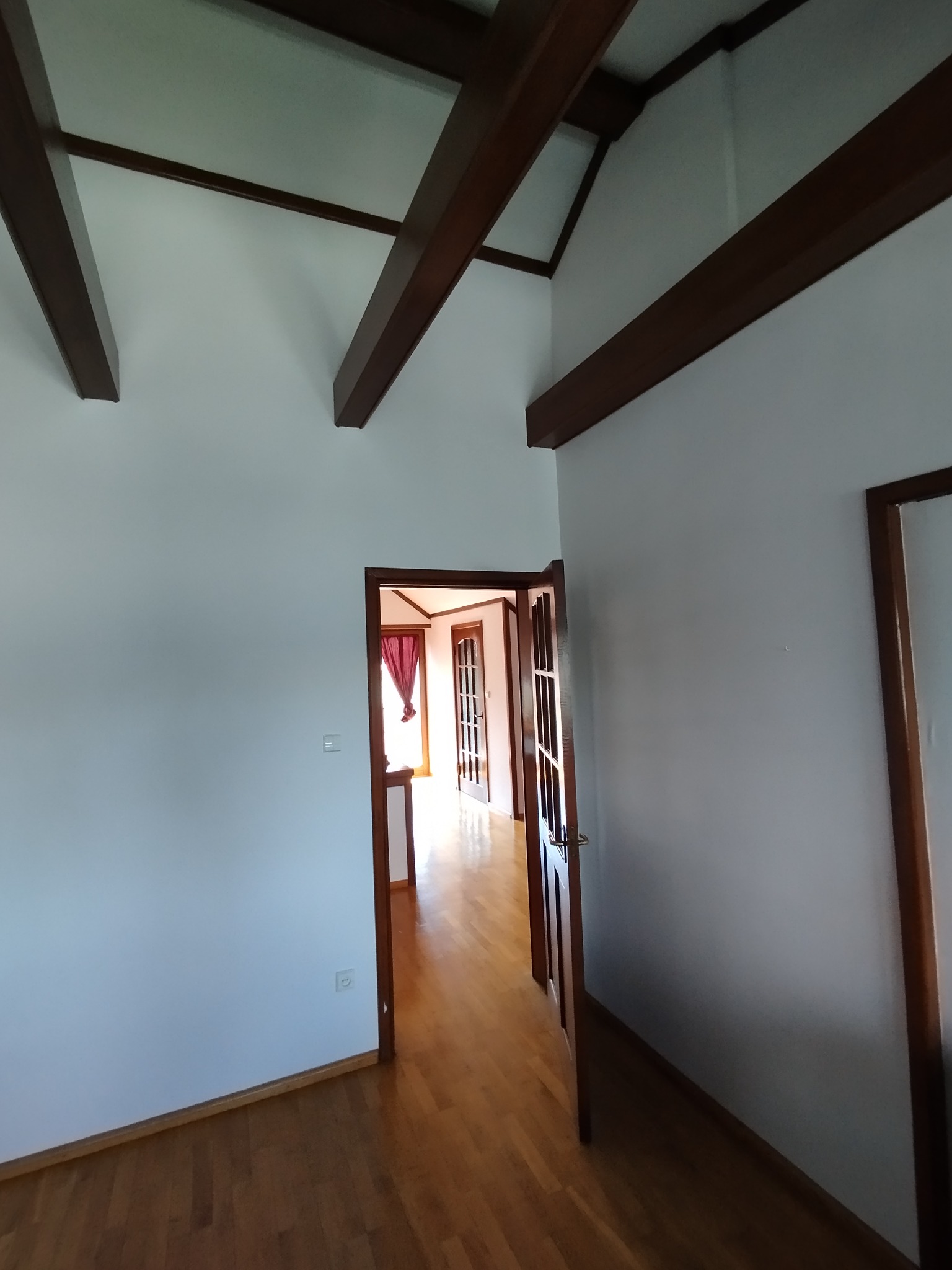 Wnętrze z białymi ścianami, drewnianymi belkami stropowymi i podłogą, widok na otwarte drzwi do kolejnego pomieszczenia z czerwonymi zasłonami.