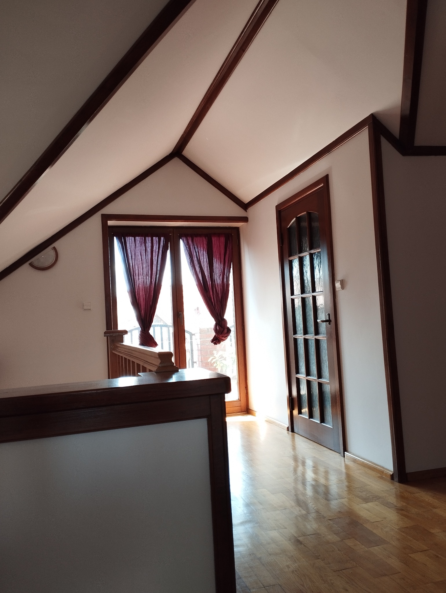 Poddasze z drewnianymi belkami, oknem balkonowym z bordowymi zasłonami i przeszklonymi drzwiami, świeżo po remoncie, widoczna czysta, pomalowana na biało skośna ściana.