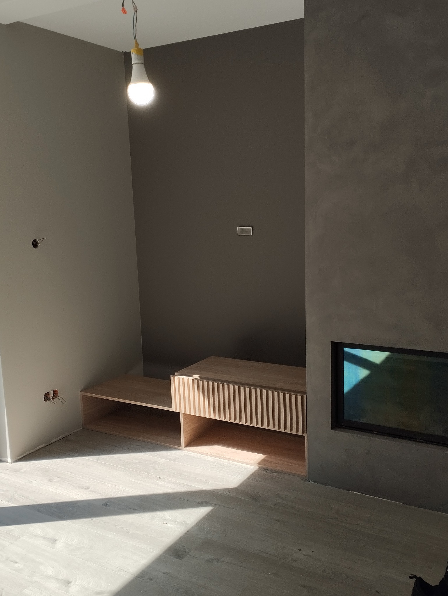 Surowe wnętrze w odcieniach szarości z drewnianą zabudową pod telewizor z lamelami, widoczne niedokończone prace elektryczne i surowy tynk dekoracyjny.