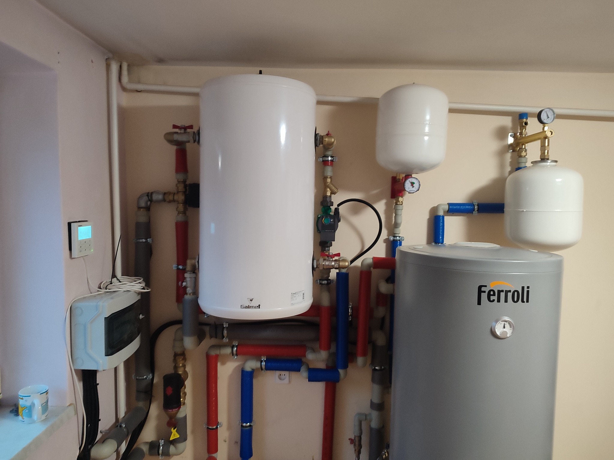 Instalacja centralnego ogrzewania z zbiornikami Galmet i Ferroli, rurami w kolorach czerwonym i niebieskim, oraz elementami sterującymi na ścianie.