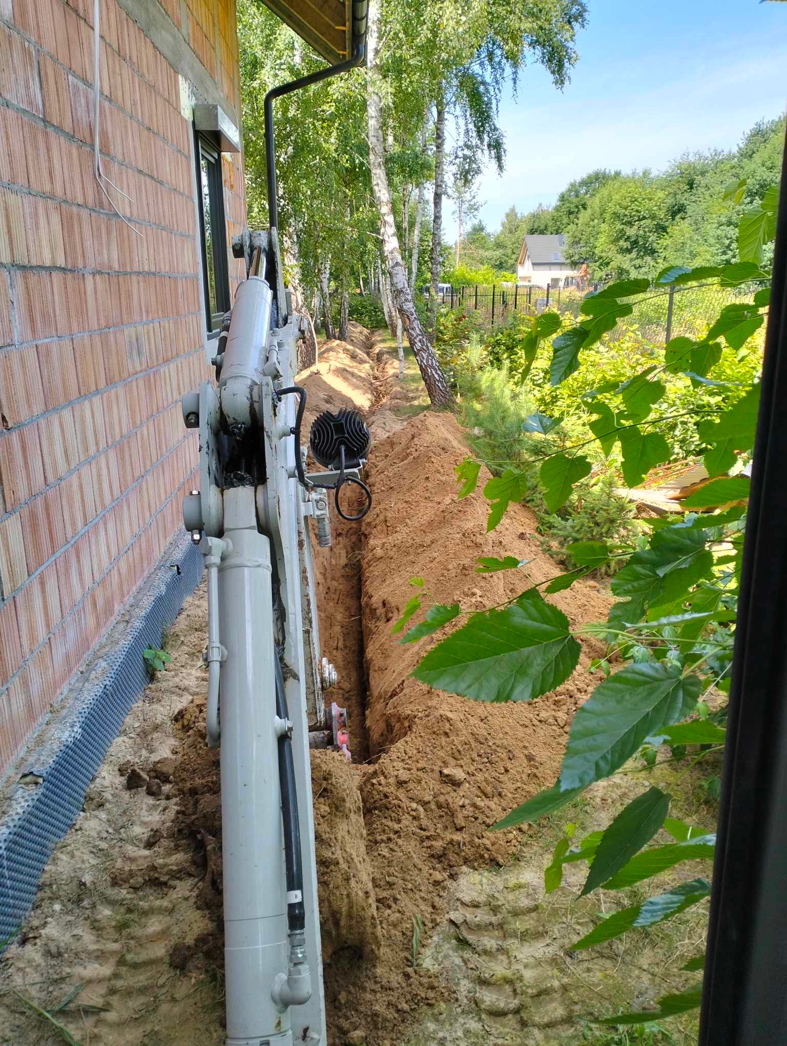 Mini koparka wykopuje wąski rów wzdłuż ściany budynku z czerwonej cegły, widoczna folia fundamentowa, w tle drzewa i ogrodzenie.