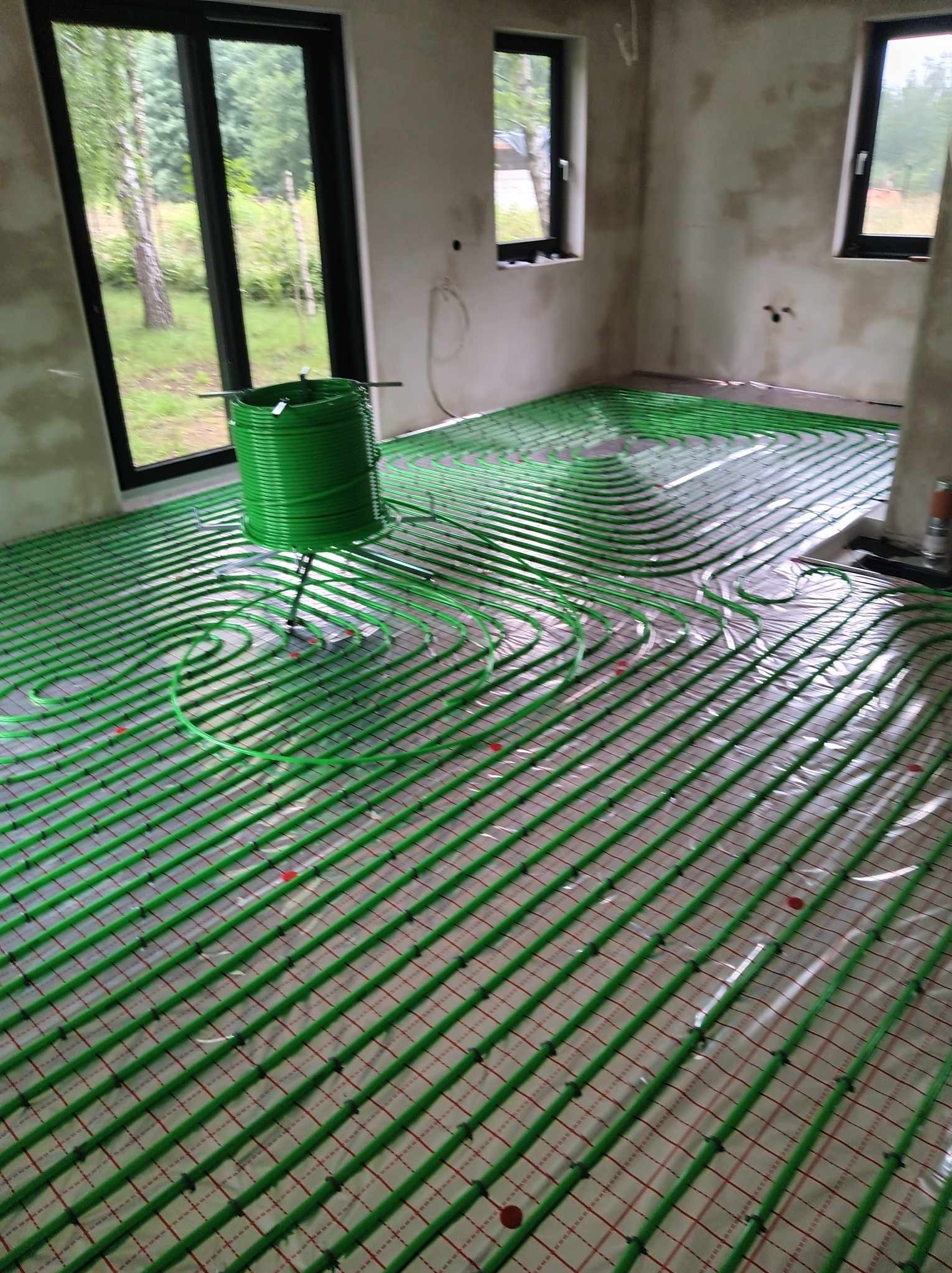 Wnętrze w stanie surowym z rozłożoną instalacją ogrzewania podłogowego z zielonych rurek na folii z siatką, widoczny zwój rurek na stojaku, okna z widokiem na zewnątrz.