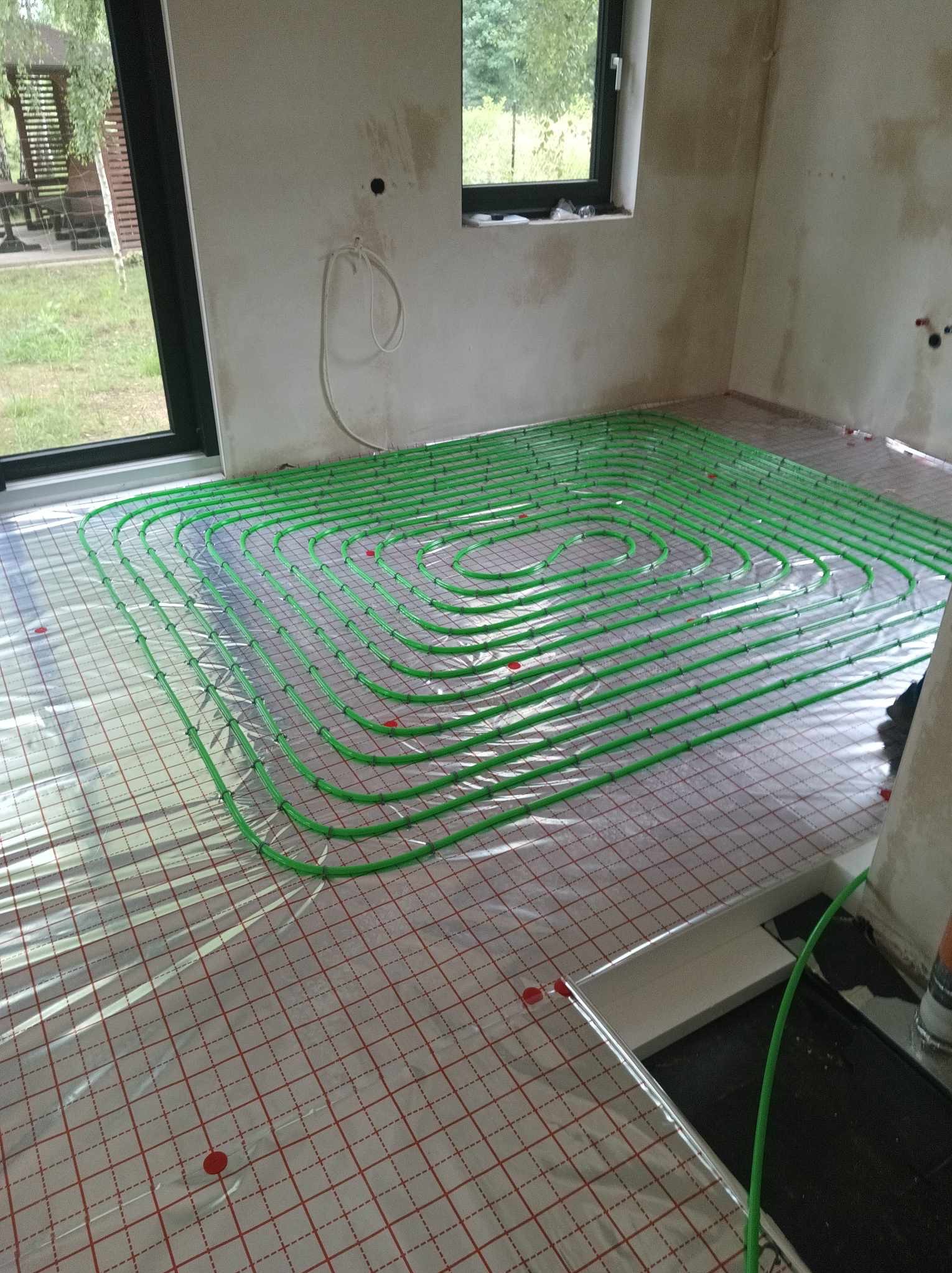 Wnętrze w trakcie montażu ogrzewania podłogowego: zielone rurki ułożone spiralnie na folii z siatką w kratkę, widoczne okno i fragment ściany w stanie surowym.