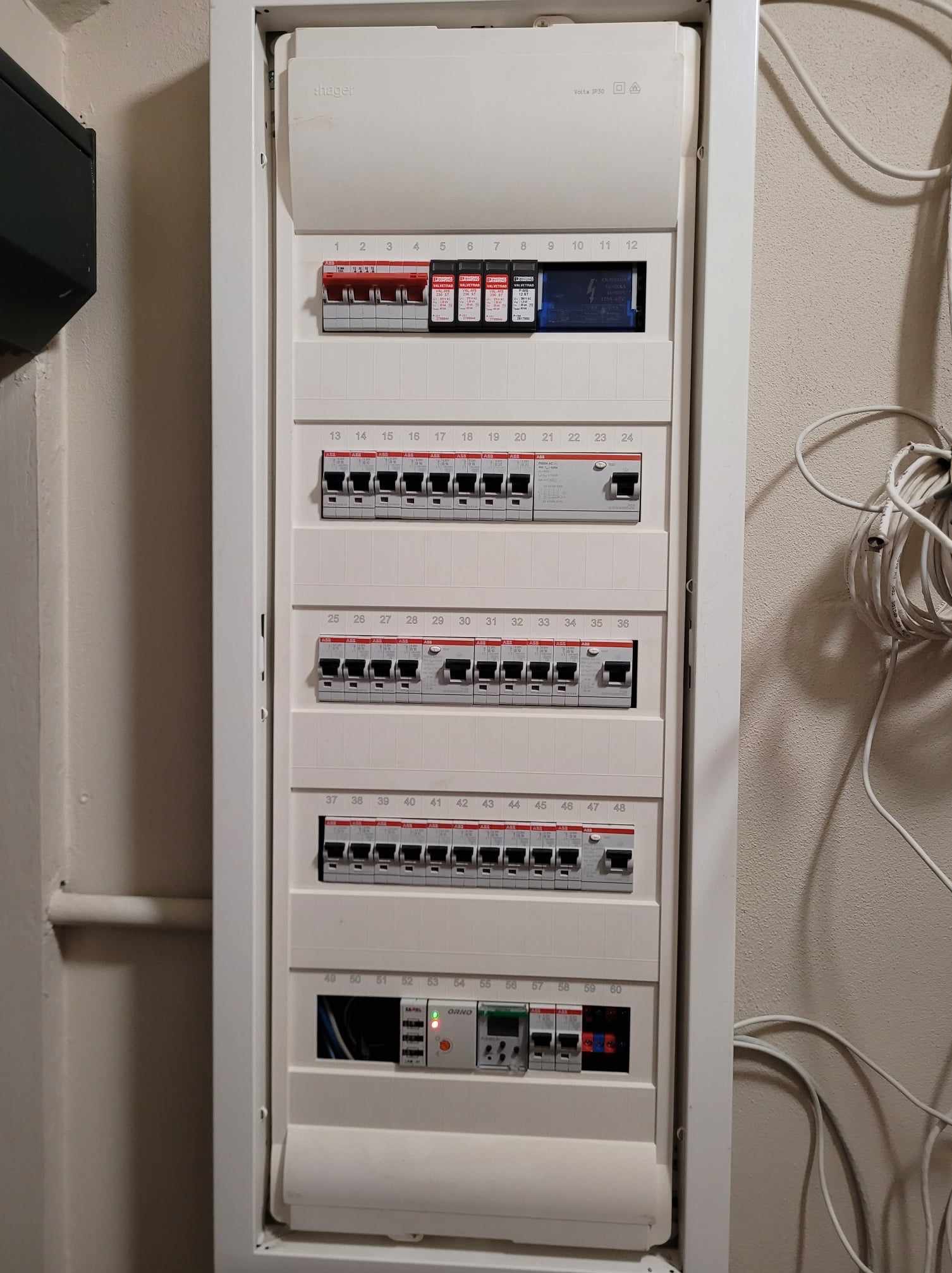 Rozdzielnica elektryczna w białej obudowie, widoczne bezpieczniki i przewody, numeracja od 1 do 60, zamontowana na ścianie.