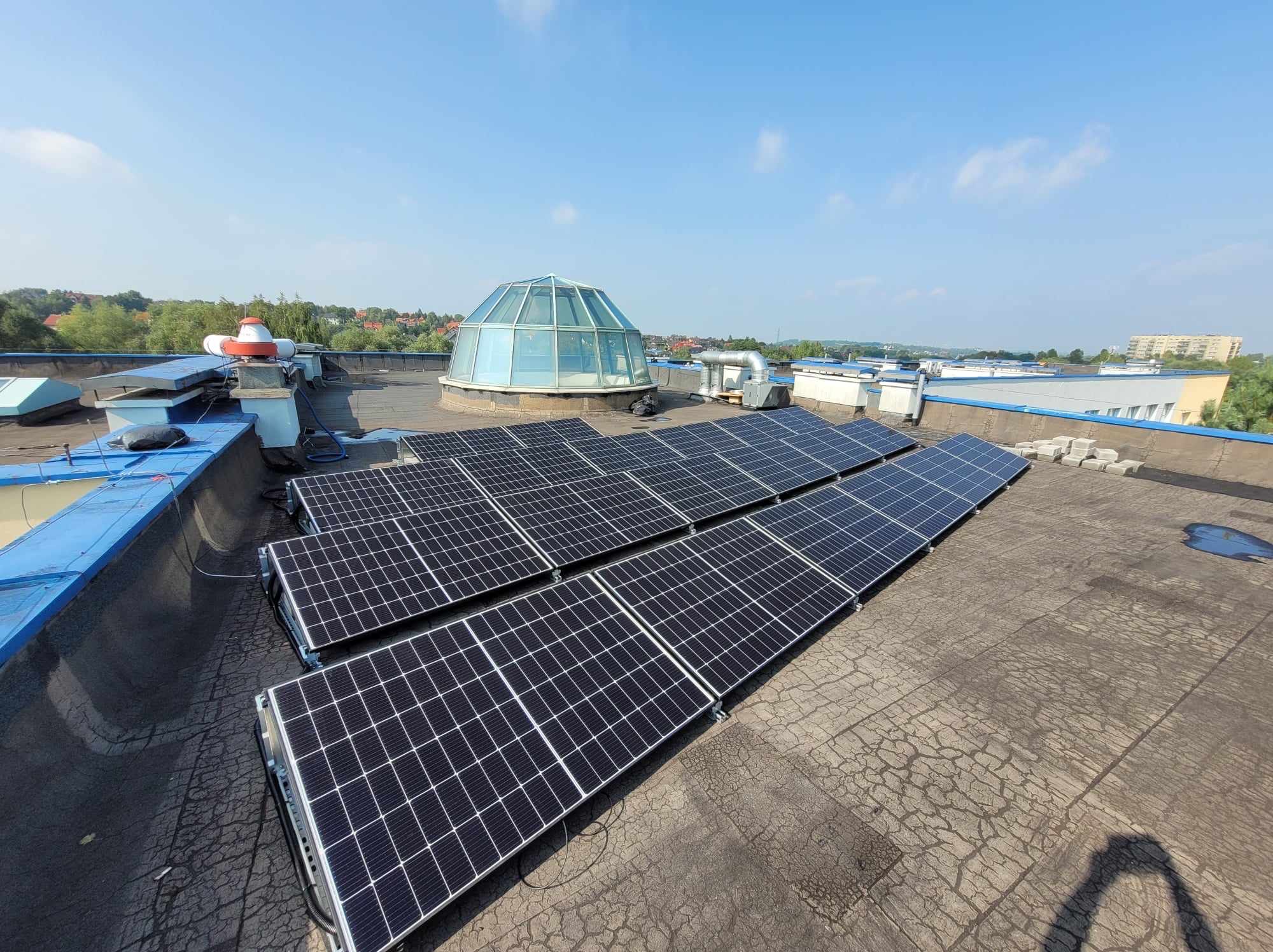 Rząd paneli fotowoltaicznych na płaskim dachu budynku z widoczną kopułą świetlika i elementami infrastruktury, w tle zabudowa miejska i błękitne niebo.