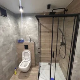 Nowoczesna łazienka z kabiną prysznicową typu walk-in z czarnymi profilami, podwieszaną toaletą i płytkami imitującymi drewno na ścianach i podłodze. Widoczna butelka płynu do czyszczenia obok...