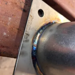 Szlifowany spaw łączący rurę ze stalową płytą, widoczne ślady obróbki i przebarwienia termiczne.