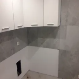 Fragment kuchni po remoncie: białe szafki wiszące z prostymi uchwytami, ściana wyłożona szarymi płytkami imitującymi marmur, widoczne gniazdko elektryczne i otwór instalacyjny.