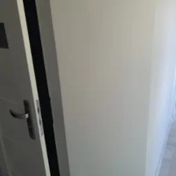 Białe, częściowo otwarte drzwi z metalową klamką, przylegające do świeżo pomalowanej, białej ściany. W tle widoczne wiaderka z farbą i folia ochronna na podłodze.