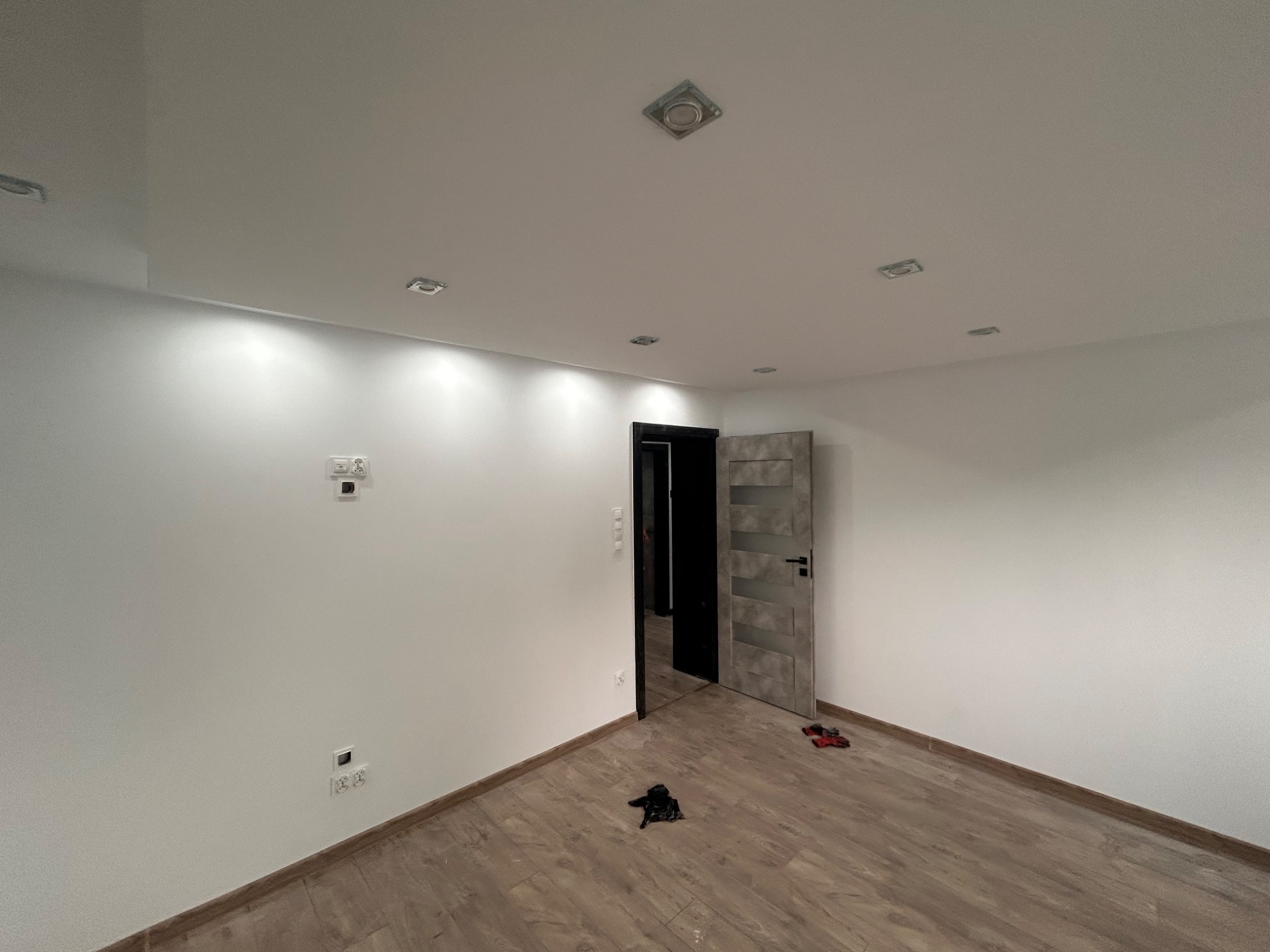 Pusty pokój po remoncie z drewnopodobną podłogą, świeżo pomalowanymi białymi ścianami, zamontowanymi halogenami w suficie i otwartymi drzwiami z szarą okleiną.