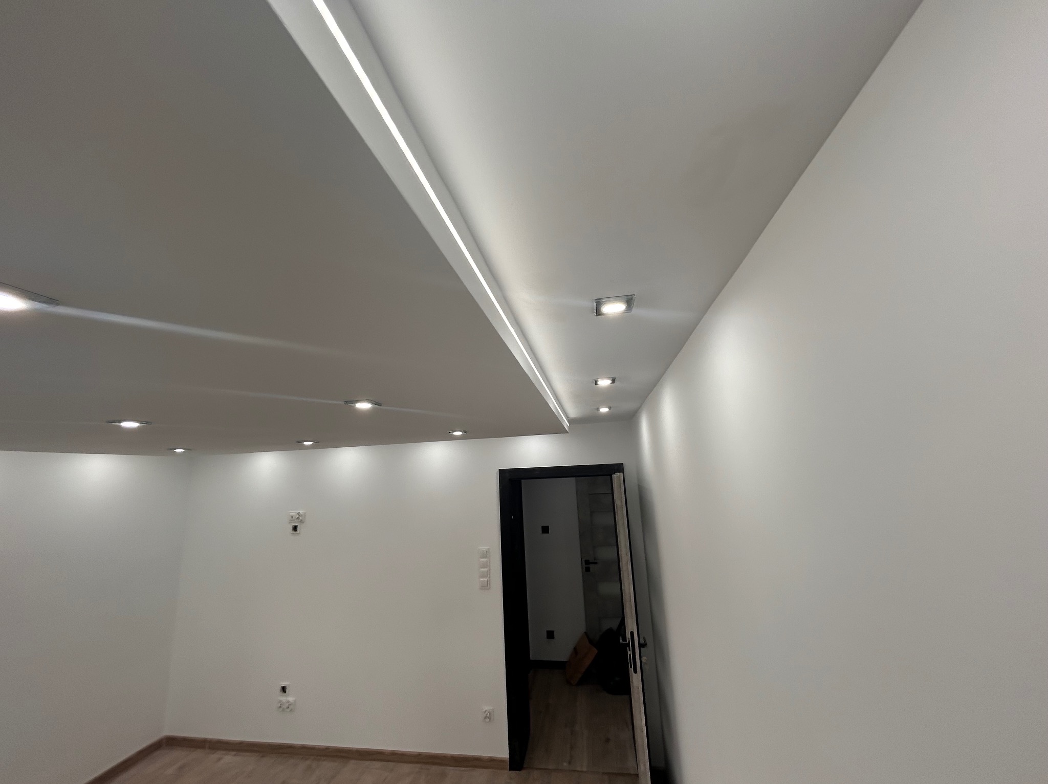 Nowoczesne wnętrze z białymi ścianami, podwieszanym sufitem z liniowym oświetleniem LED i punktowymi lampami, widok na otwarte drzwi do kolejnego pomieszczenia.