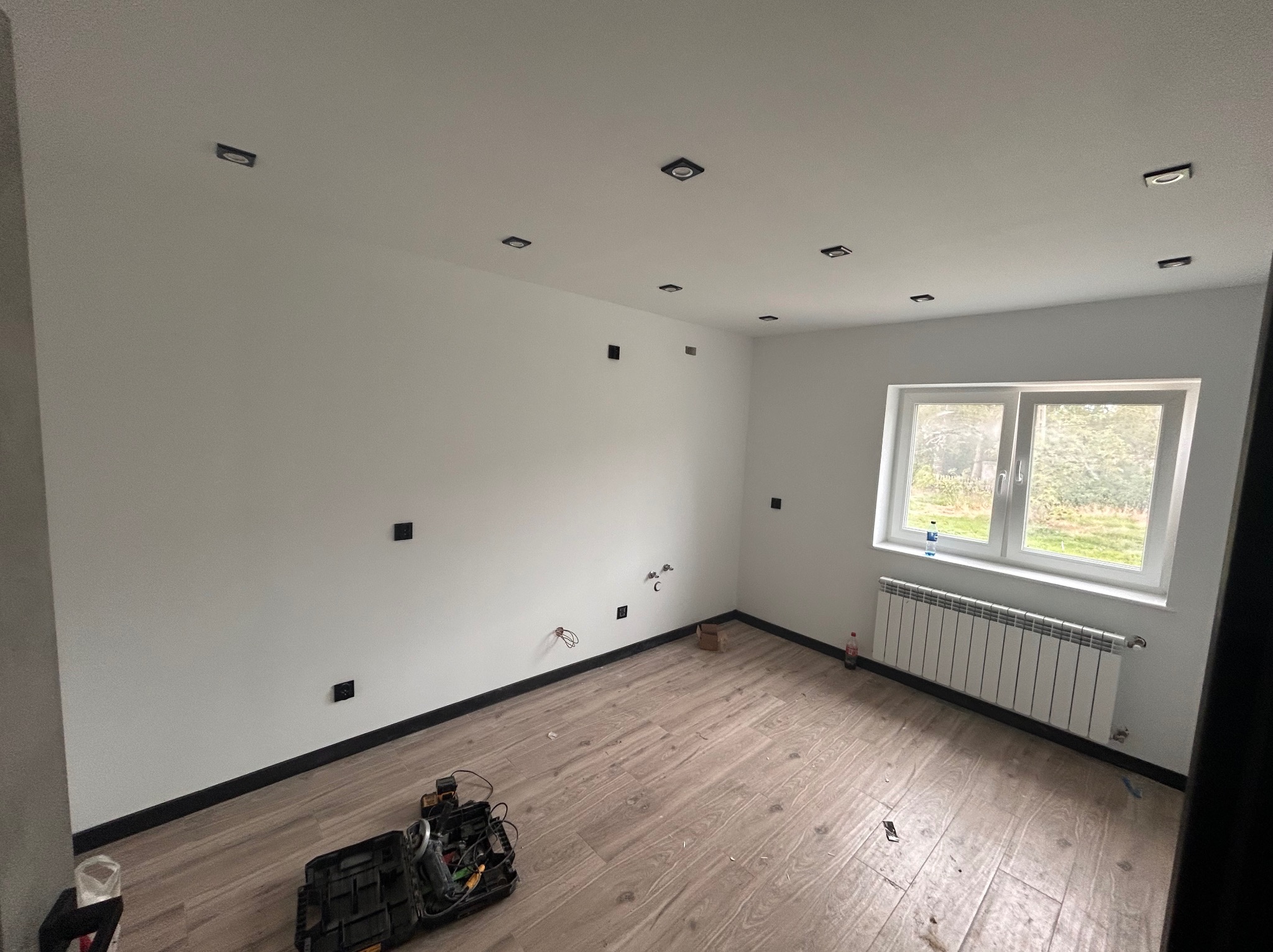 Pusty pokój po remoncie, z nową podłogą, świeżo pomalowanymi ścianami z przygotowanymi punktami oświetleniowymi i gniazdkami, grzejnik pod oknem, skrzynka z narzędziami na podłodze.