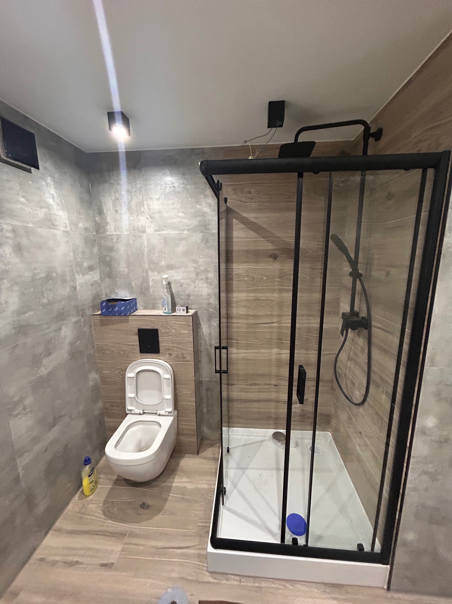 Nowoczesna łazienka z kabiną prysznicową typu walk-in z czarnymi profilami, podwieszaną toaletą i płytkami imitującymi drewno na ścianach i podłodze. Widoczna butelka płynu do czyszczenia obok...