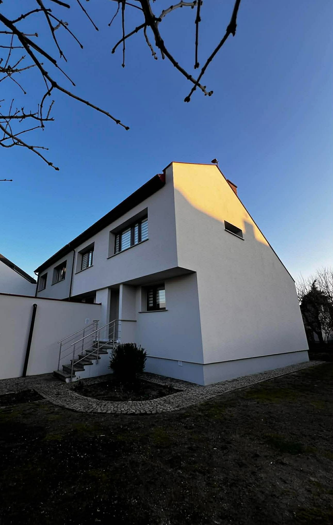 Biały, nowoczesny dom jednorodzinny z oknami i schodami wejściowymi, oświetlony popołudniowym słońcem, na tle błękitnego nieba i gałęzi drzew.