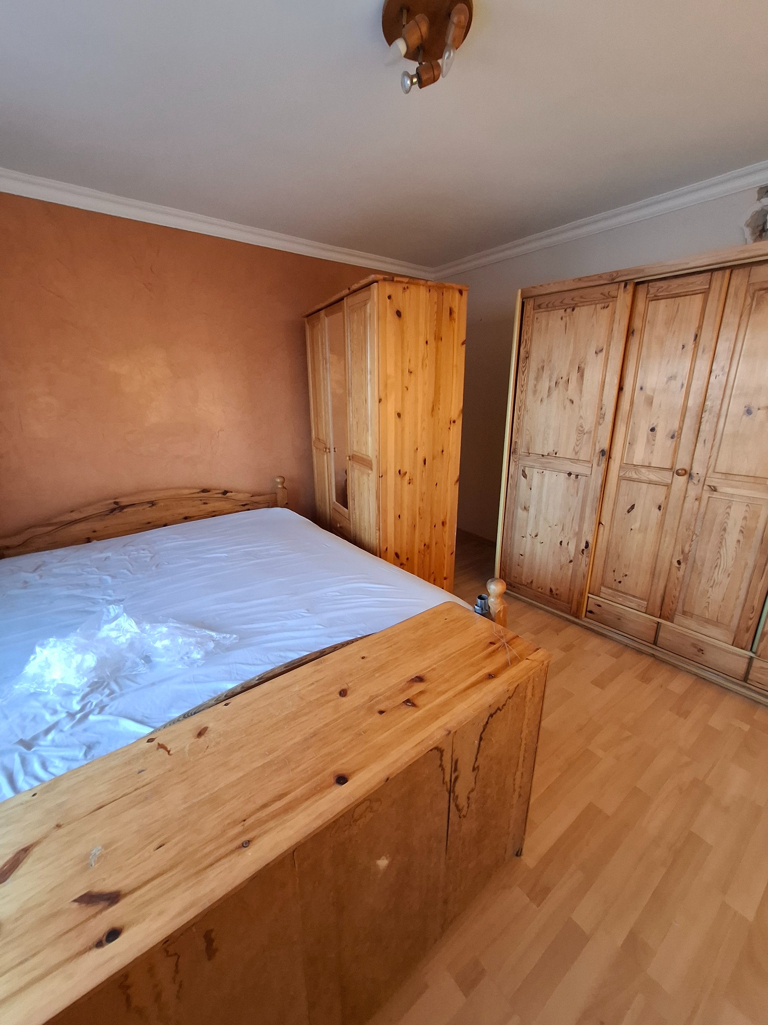 Rustykalna sypialnia z drewnianym łóżkiem, szafami i pomarańczową ścianą. Widoczne detale wykończenia i oświetlenie sufitowe. Podłoga z paneli.